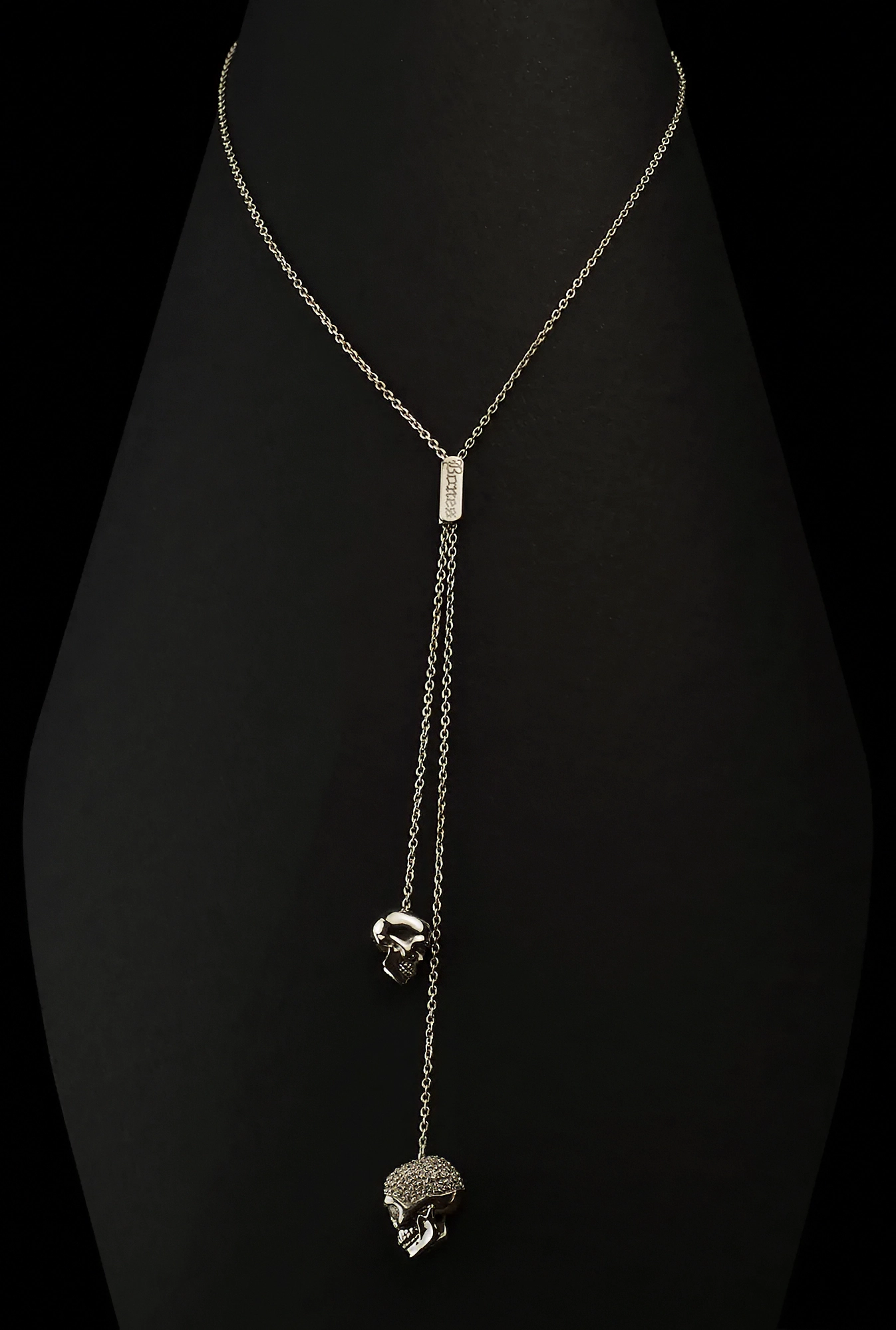 Low key still life photography of silver skull double pendants by Bones Firenze on a black background – dramatic lighting, minimalist composition