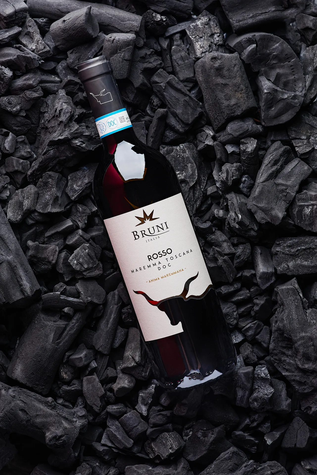 Ambient still life photo of Maremma Toscana DOC wine bottle lying on charcoal – elegant Tuscan wine product photography